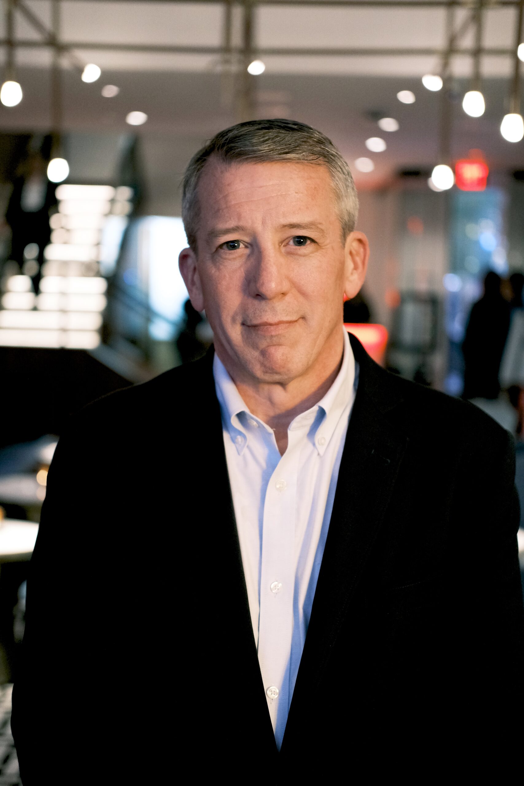 Headshot of Dan Ronayne standing in a dress shirt and coat with lights in the background.