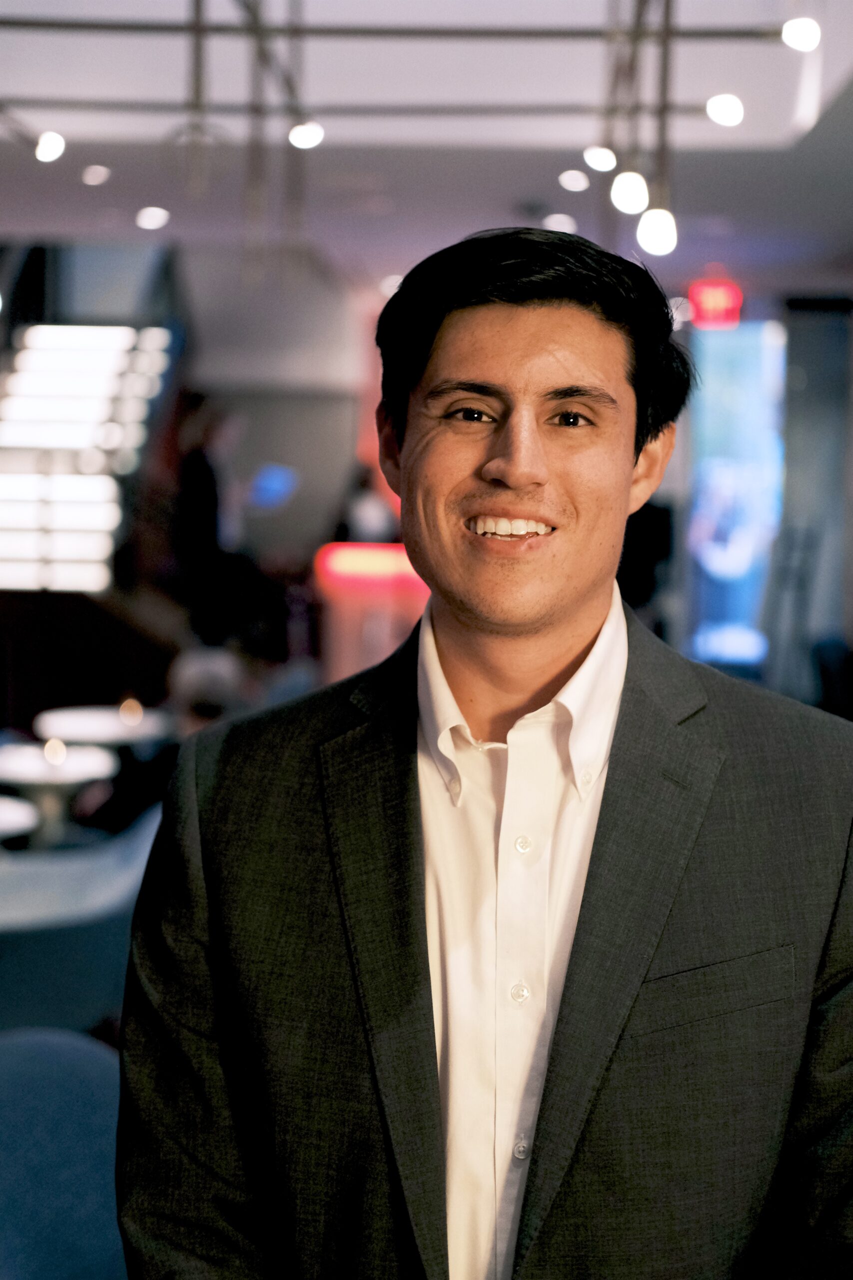 Headshot of Brendan Montesinos standing in a dress shirt and coat with lights in the background.
