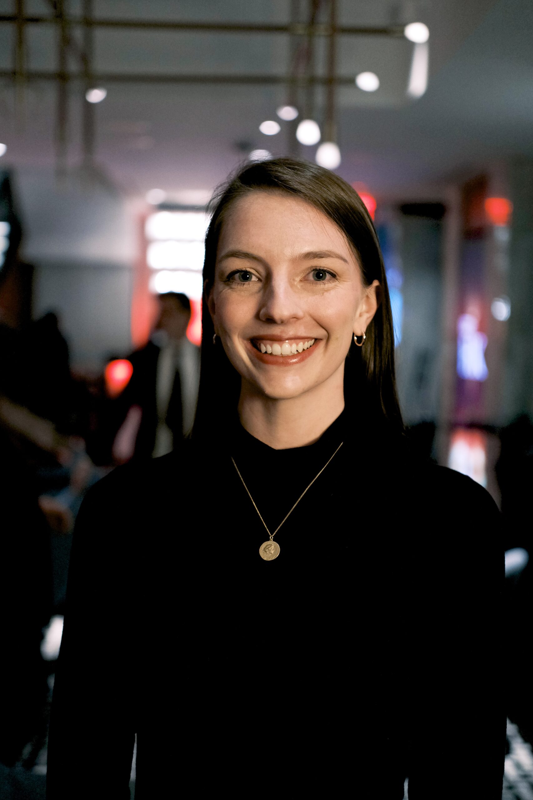 Headshot of Kirby Eule standing wearing black shirt and necklace with lights in the background.